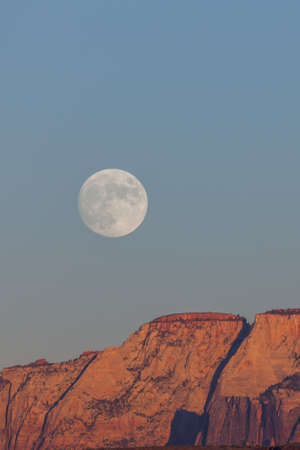 The full moon rising over the dramatic mountains of Zion National Park lit by the setting sun with a clear sky.の写真素材