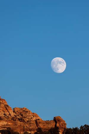 An almost full moon rising above red sandstone mountains with clear a blue evening sky near Zion National Park, Utah.の写真素材
