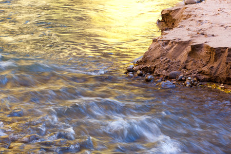The Virgin River flowing over river rocks next to a sandy embankment with a yellow glow on the surface from fall trees reflected in the sunshine at Zion National Park, Utah.の写真素材