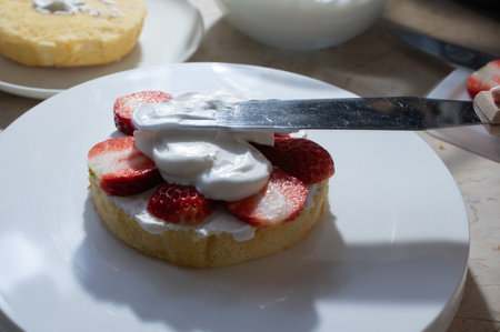 A sponge cake sheet with whipped cream and sliced strawberries being appliedの写真素材