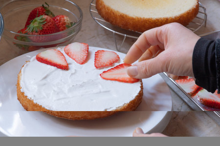 A sponge cake sheet with whipped cream applied and sliced strawberries being placed by human hand on a marble table surrounded by ingredients.の写真素材