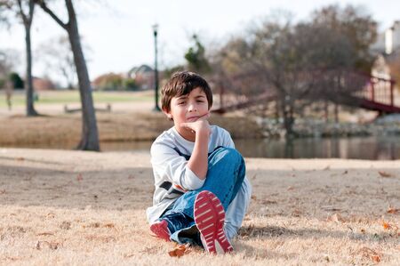 Cute little boy sitting in grass during the fall at a park with pond and bridge in background.