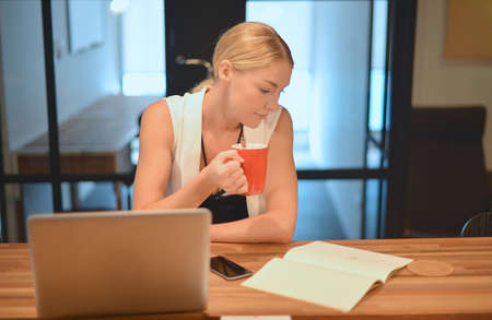 Business blonde girl drinking a cup of coffee in front of a laptopの写真素材