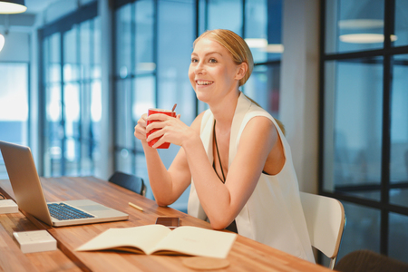 Business blonde girl drinking a cup of coffee in front of a laptopの写真素材