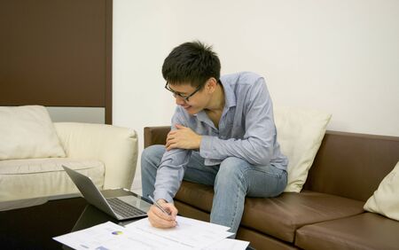 Asian businessman writing a document with a laptop in living roomの写真素材