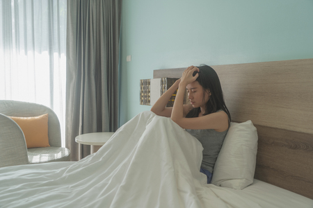 Asian woman thinking about problems and suffering from depression on bed in a modern bedroom with white blanket.の写真素材