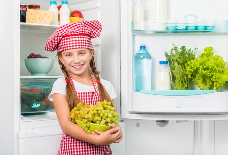little cook girl holding grapes near opened fridgeの写真素材