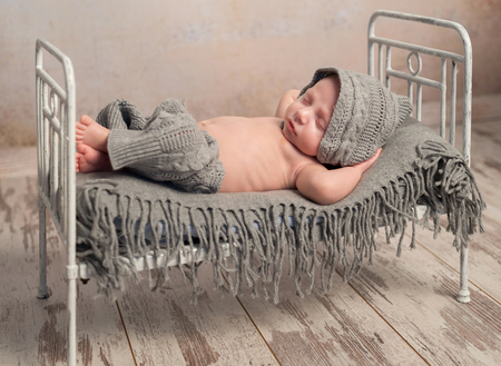 funny newborn baby in knitted hat and pants sleeping on old retro cot with plaid with hands behind headの写真素材