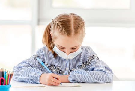 Schoolgirl in protective mask writing in notebook