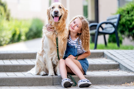 Preteen girl wearing hat hugging golden retriever dog sitting outdoors in summertime. Pretty kid petting fluffy doggy pet in cityの写真素材