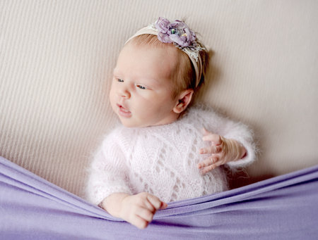 Newborn baby girl wearing flower wreath and knitted sweater lying and looking back with mouth open. Sweet infant child kid studio portraitの写真素材