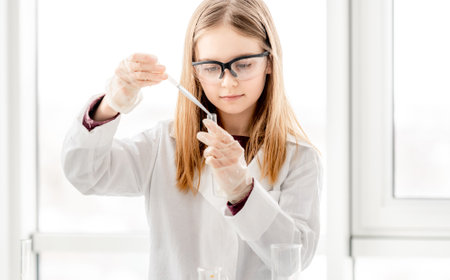 Smart girl during scientific chemistry experiment wearing protection glasses, holding tubes and measuring ingridients. Schoolgirl with chemical equipment on school lessonの写真素材