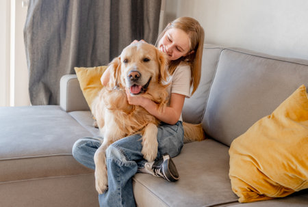 Preteen girl hugging golden retriever dog and smiling sitting on sofa at home. Beautiful child kid with labrador doggy pet indoorsの写真素材
