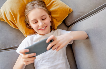 Preteen girl with smartphone chatting with friends lying on sofa at home. Pretty child kid smiling with modern cellphone in social media on couchの写真素材