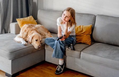 Preteen girl with golden retriever dog holding tv remote control and sitting on sofa at home. Beautiful child with labrador doggy pet watching cartoons at television kids channelの写真素材