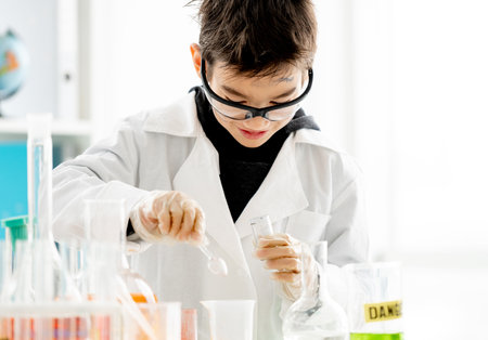School boy wearing protection glasses with dirty face doing chemistry experiment in elementary science class and measuring components with spoon. Clever pupil in labの写真素材