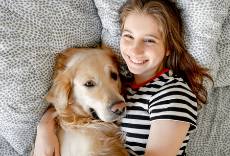 Pretty girl hugging golden retriever dog and smiling lying in bed. Happy teenager with purebred doggy pet labrador resting togetherの写真素材