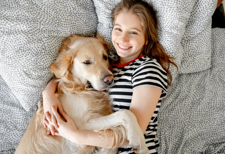 Pretty girl hugging golden retriever dog and smiling lying in bed. Happy teenager with purebred doggy pet labrador resting togetherの写真素材