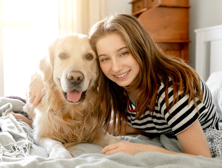 Pretty girl hugging golden retriever dog and smiling sitting in bed. Female teenager with purebred doggy pet labrador looking at camera at homeの写真素材