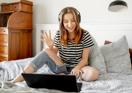 Pretty girl with headphones and laptop in bed during online learning webinar. Beautiful teenager talking by video call with pc computer at bedroomの写真素材
