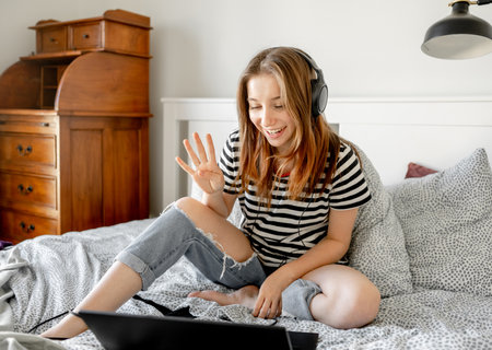 Pretty girl with headphones and laptop in bed during online learning webinar. Beautiful teenager talking by video call with pc computer at bedroomの写真素材