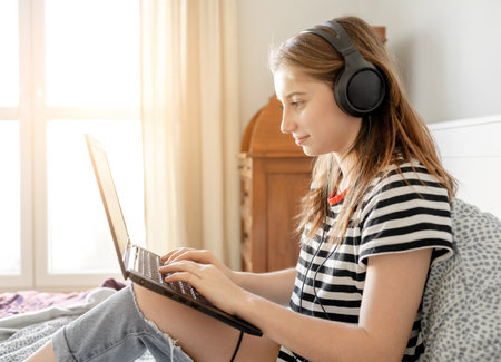 Pretty girl with headphones and laptop in bed listening online webinar. Beautiful teenager making video call with computer at bedroomの写真素材