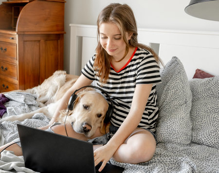 Pretty girl with golden retriever dog, headphones and laptop in bed. Female teenager with purebred doggy pet labrador making video call at pc onlineの写真素材