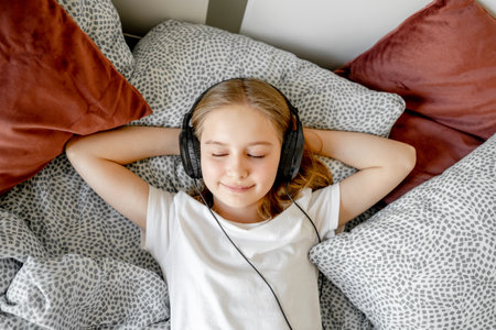 Pretty girl child in headphones lying in bed and listening music at home. Beautiful female kid resting with earpods in bedroom and smilingの写真素材