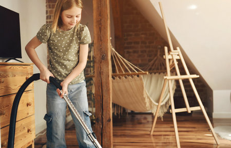 Girl child cleaning carpet with vacuum cleaner from dust at home. Preteen female kid with hoover making order in apartmentの写真素材