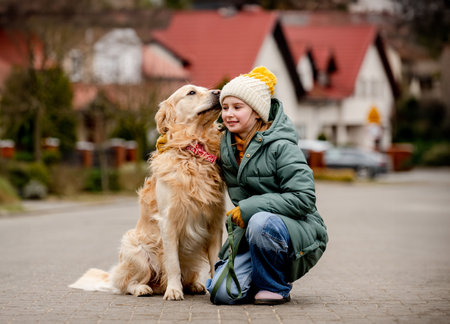 Preteen child girl sitting with golden retriever dog at autumn city street wearing hat and warm jacket.の写真素材