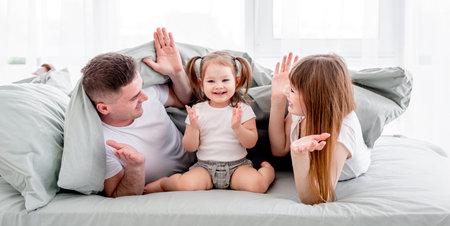 Beautiful family under the blanket in the bed holding hands up and smiling. Young parents playing with small daughter under coverlet.の写真素材