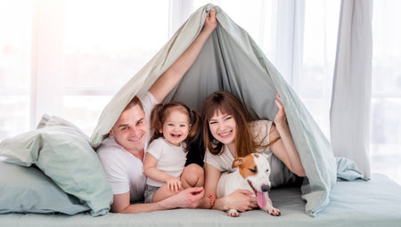 Smiling family with dog under the blanket in the bed. Beautiful morning moments of mother, father and little daughter with pet under coverlet.の写真素材
