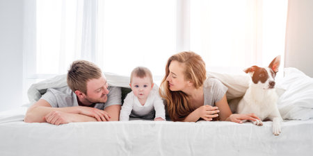 Mother and father lying in the bed under blanket with their son and cute dog. Beautiful family morning together with pet. Happy parenthood momentsの写真素材