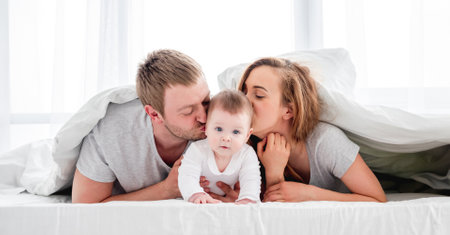 Mother and father lying in the bed under blanket with their son and kissing baby on his cheeks. Beautiful family morning together in sunny bedroom.の写真素材