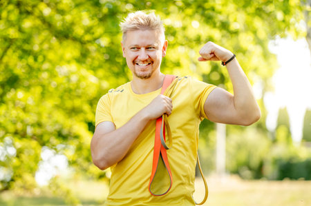 Man with elastic rubber band showing muscles outdoors after arm workout. Guy with sport equipment looking at camera and smilingの写真素材