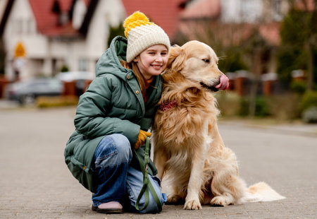 Preteen child girl sitting with golden retriever dog at autumn city street wearing hat and warm jacket. Pretty kid hugging purebred pet doggy labrador outdoorsの写真素材