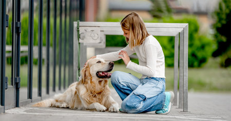 Preteen girl with golden retriever dog sitting in city at street. Pretty child kid petting purebred pet doggy outdoorsの写真素材