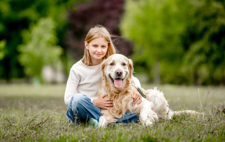 Preteen girl with golden retriever dog sitting at nature and looking back. Cute child kid hugging purebred pet doggy in park at summerの写真素材