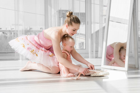 Two ballerinas, mother and daughter sitting on the twine in front of large windows in studio. Little ballerina girl and her teacher doing the split. Little girl stretching with her motherの写真素材