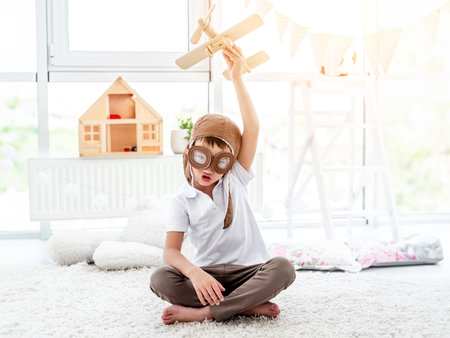 Happy little boy in pilot hat playing with wooden plane in children's roomの写真素材
