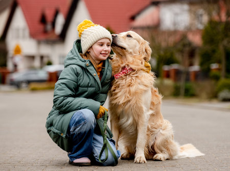 Preteen child girl sitting with golden retriever dog at autumn city street wearing hat and warm jacket. Pretty kid hugging purebred pet doggy labrador outdoorsの写真素材