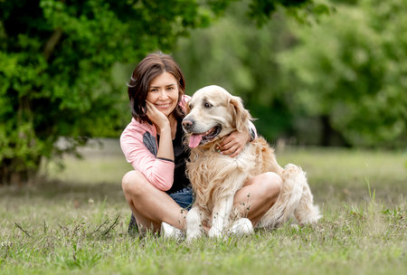 Pretty girl with golden retriever dog sitting at nature. Beautiful young woman hugging purebred pet doggy labrador in park at summerの写真素材