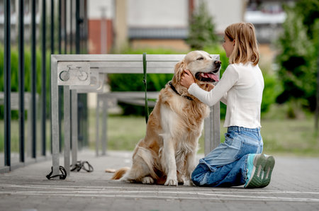 Preteen girl with golden retriever dog sitting in city at street. Pretty child kid petting purebred pet doggy outdoorsの写真素材