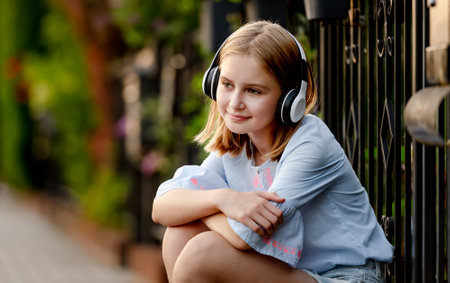 Pretty preteen child girl wearing headphones sitting near fence at city street and smiling. Cute female model with music earpods summer portraitの写真素材