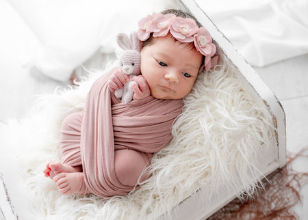Adorable newborn baby girl lying in tiny bed on fur portrait. Cute infant child kid swaddled in fabric resting in room with daylightの写真素材