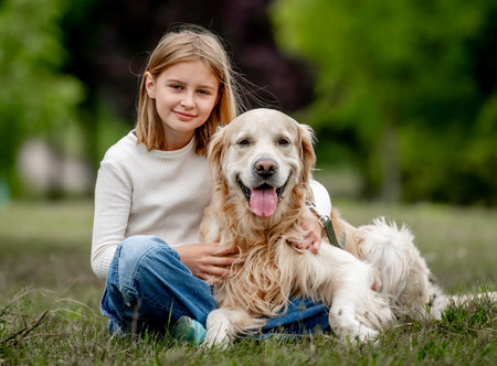 Preteen girl with golden retriever dog sitting at nature and looking back. Cute child kid hugging purebred pet doggy in park at summerの写真素材