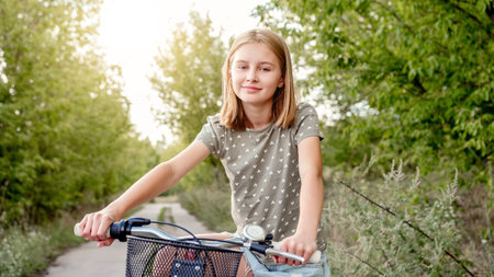 Cute preteen girl with bycicle outdoors looking at camera and smiling. Pretty child with bike at city street at summer parkの写真素材