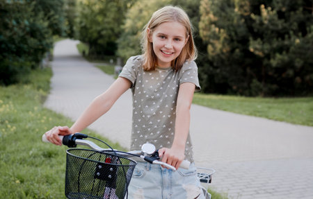 Cute preteen girl with bycicle outdoors looking at camera and smiling. Pretty child with bike at city street at summer parkの写真素材