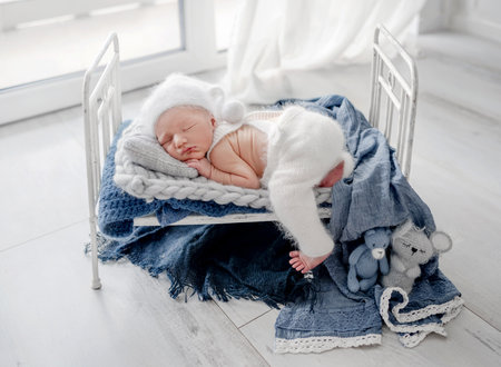 Adorable newborn baby boy sleeping in tiny bed decorated with blankets and knitted toys. Cute infant child kid napping in white warm clothesの写真素材