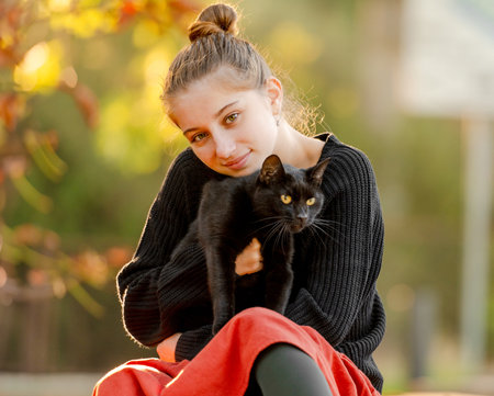 Pretty girl in red skirt hugging black cat outdoors at street with autumn leaves. Beautiful model teenager sitting with feline animal at parkの写真素材